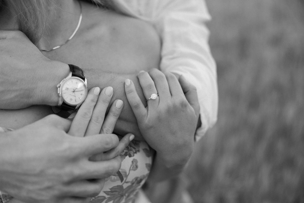 Close-up black and white photo showing hands with engagement rings against a floral dress pattern.