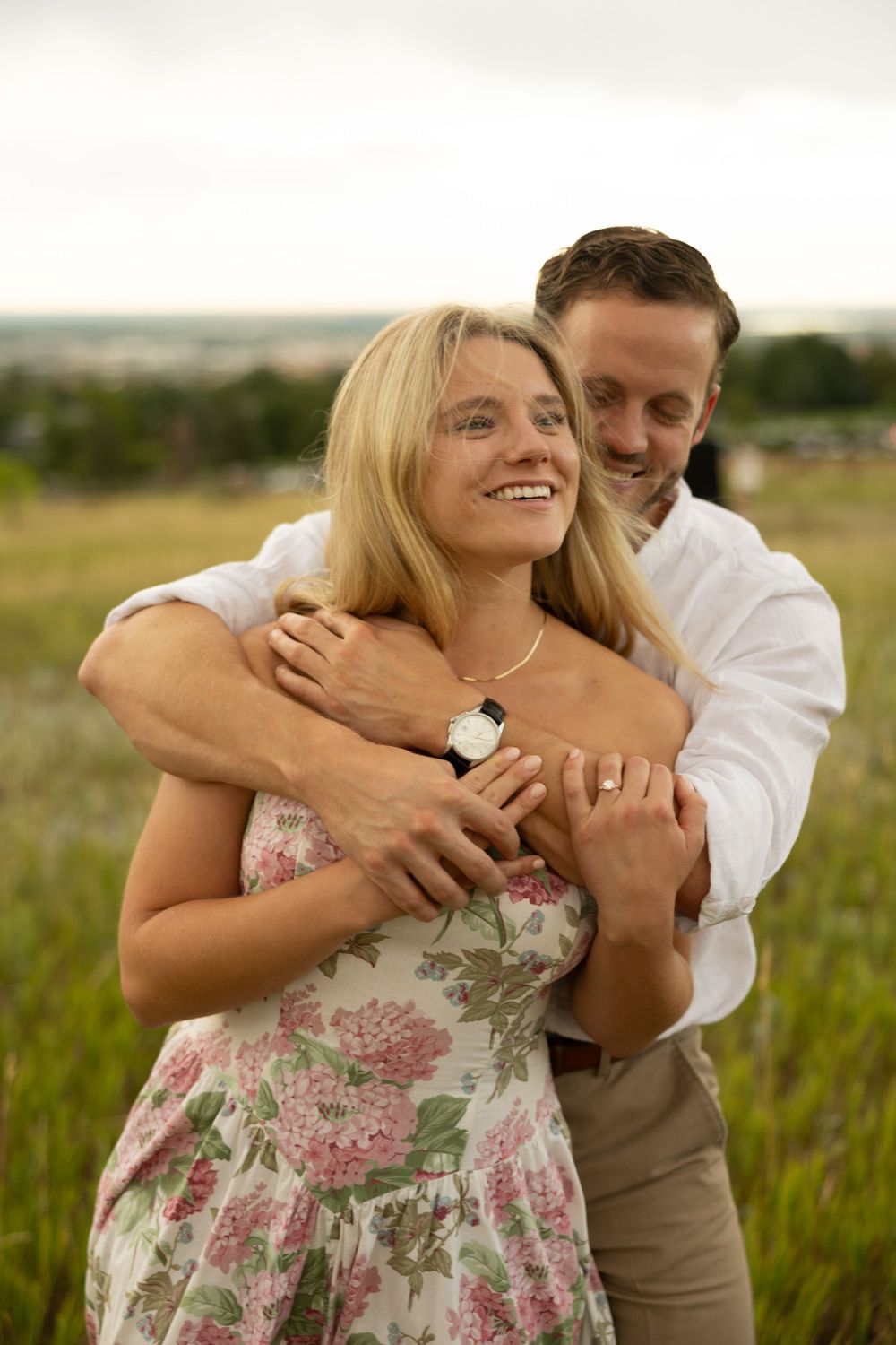 A tender embrace in a summer meadow showcasing a floral dress and engagement ring.