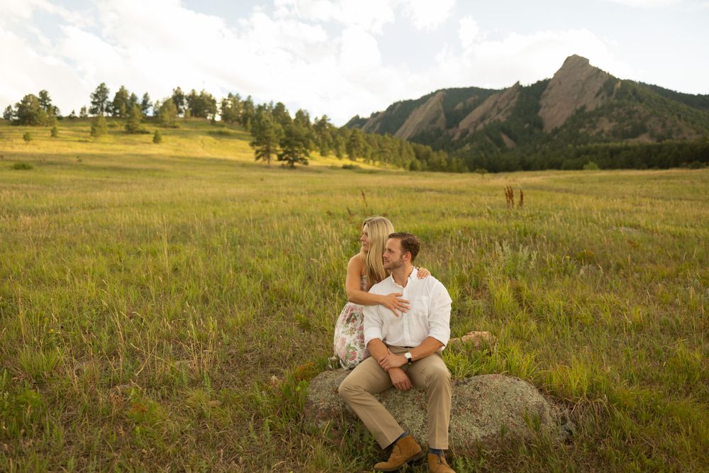 Romantic couple sits on boulder in grassy field with mountain backdrop during sunset.