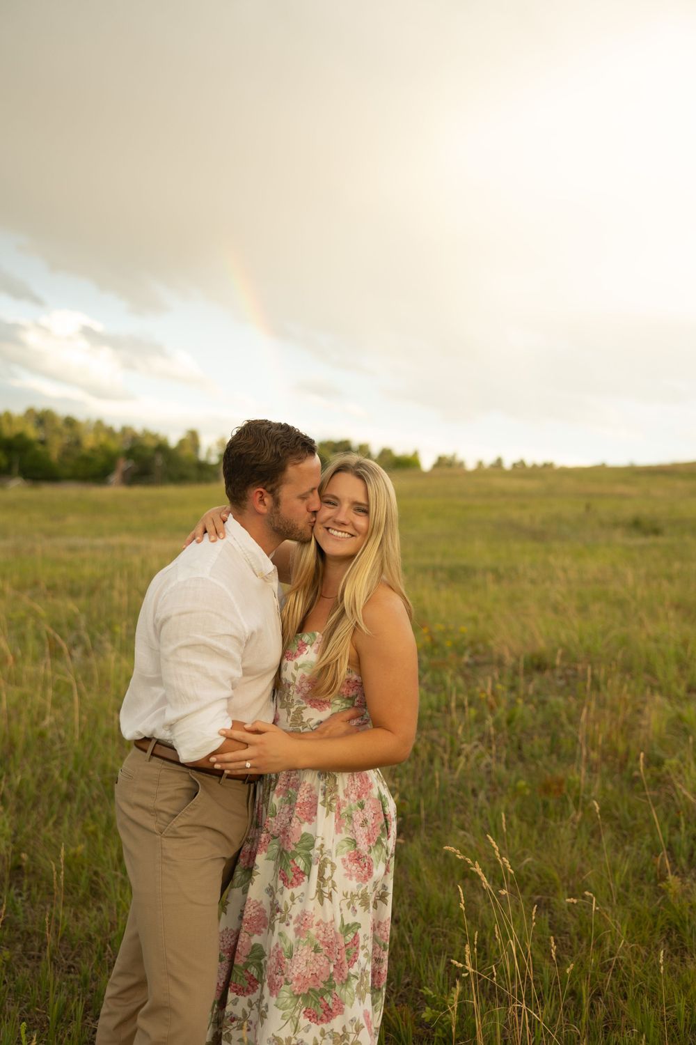 Couple embraces in floral dress and white shirt against golden mountain meadow at sunset.