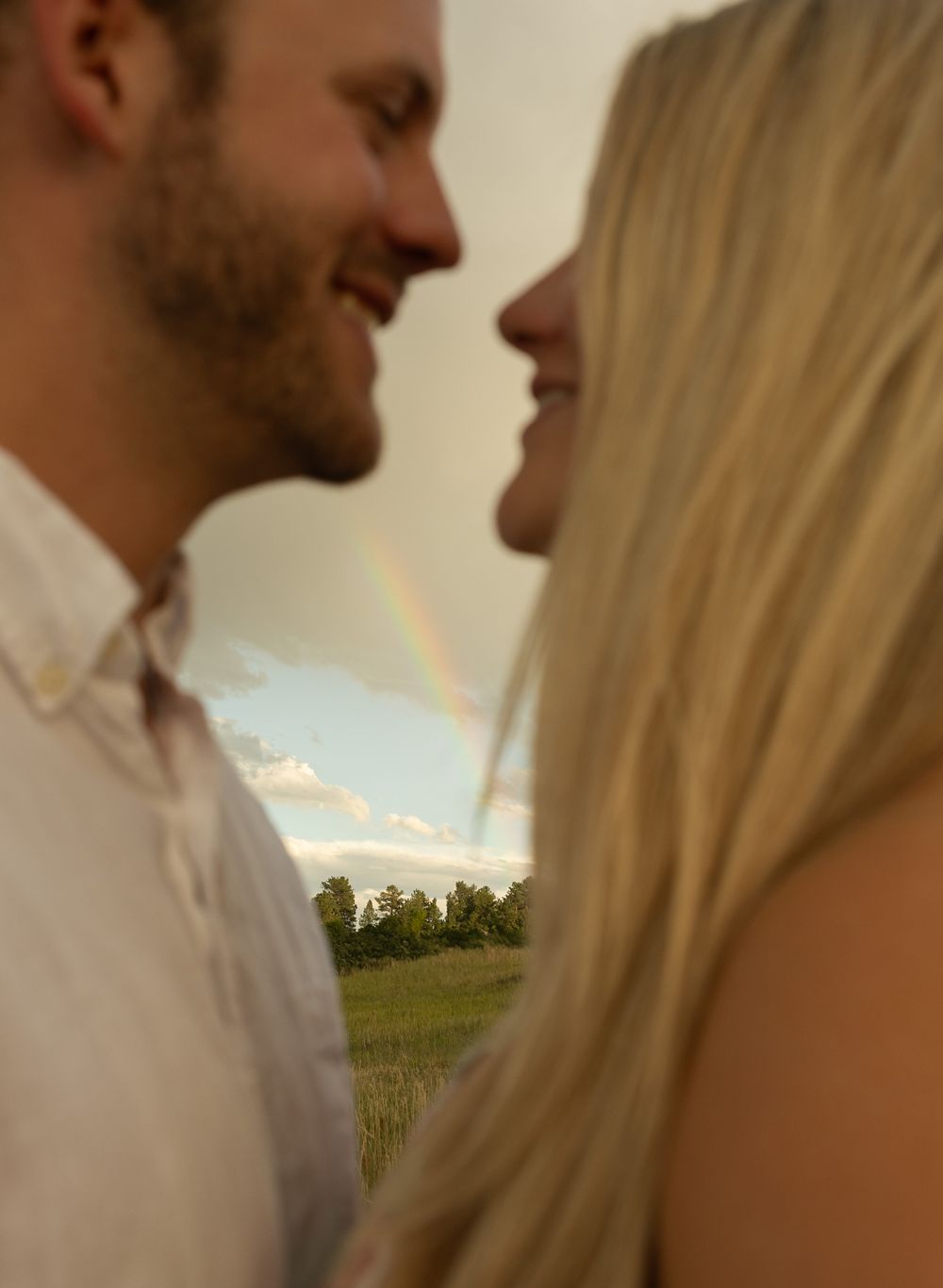 Close up moment between couple with sunlit mountain meadow and rainbow in background.