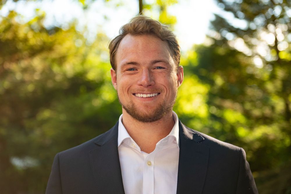 A person in a white dress shirt smiles warmly outdoors with sunlit trees and greenery in the background.