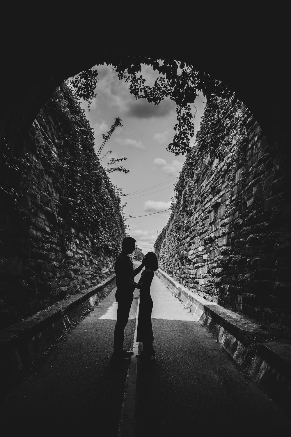 A black and white silhouette of two people standing in a stone tunnel archway under dramatic cloudy skies.
