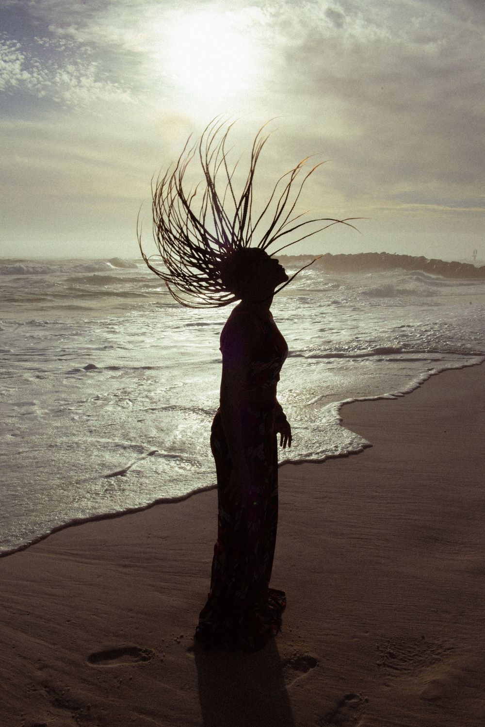 A silhouetted figure stands on a misty beach at sunset with hair flowing dramatically in the wind.