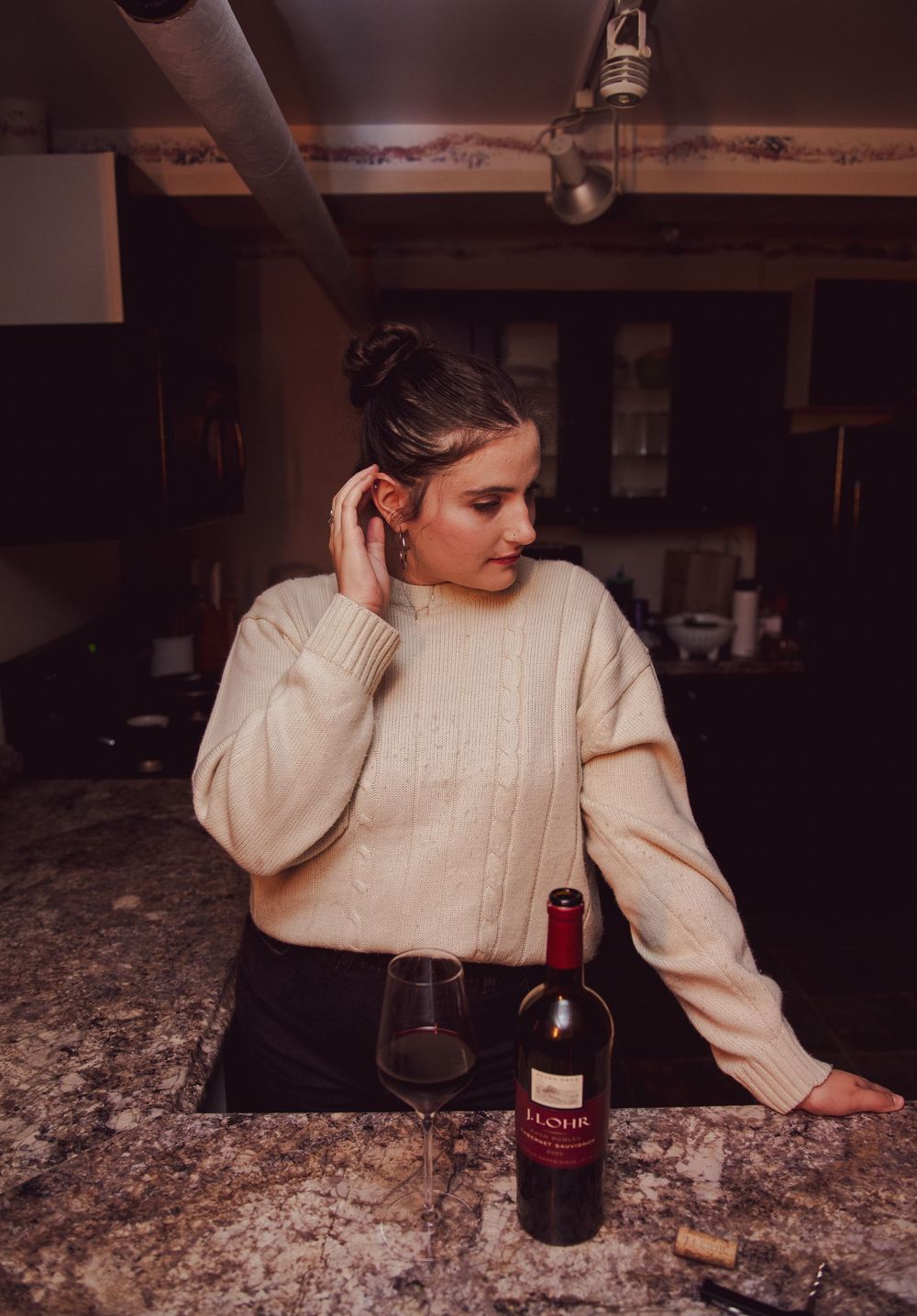 A person in a cream sweater sits at a granite countertop with a bottle of red wine in a dimly lit room.