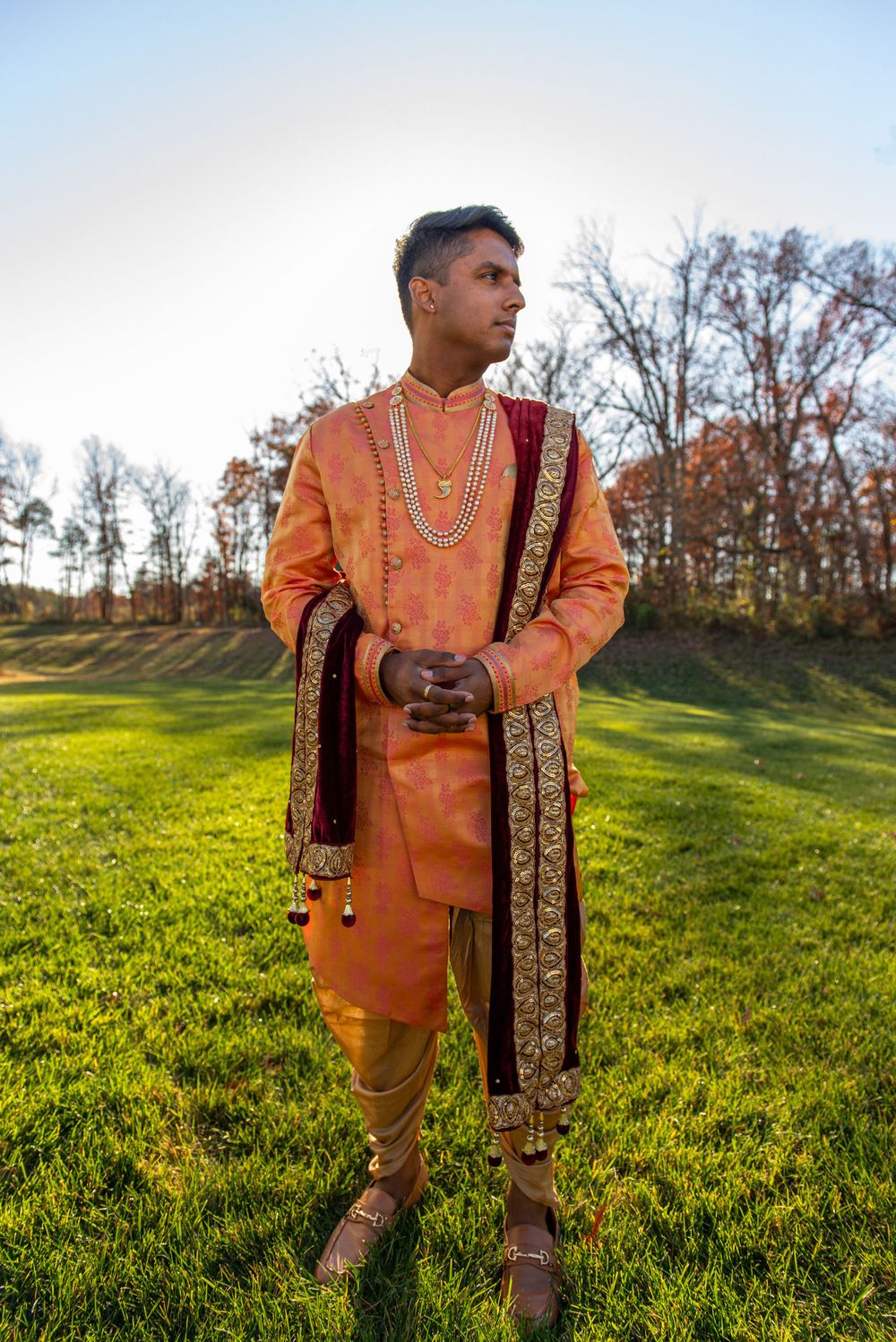 Person wearing traditional orange South Asian wedding attire with embroidered shawl poses outdoors on grassy hillside.