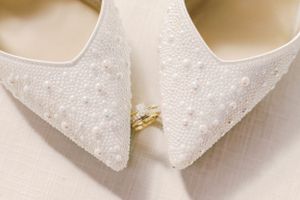 Gold wedding ring positioned between white lace bridal shoes showing elegant wedding day details.