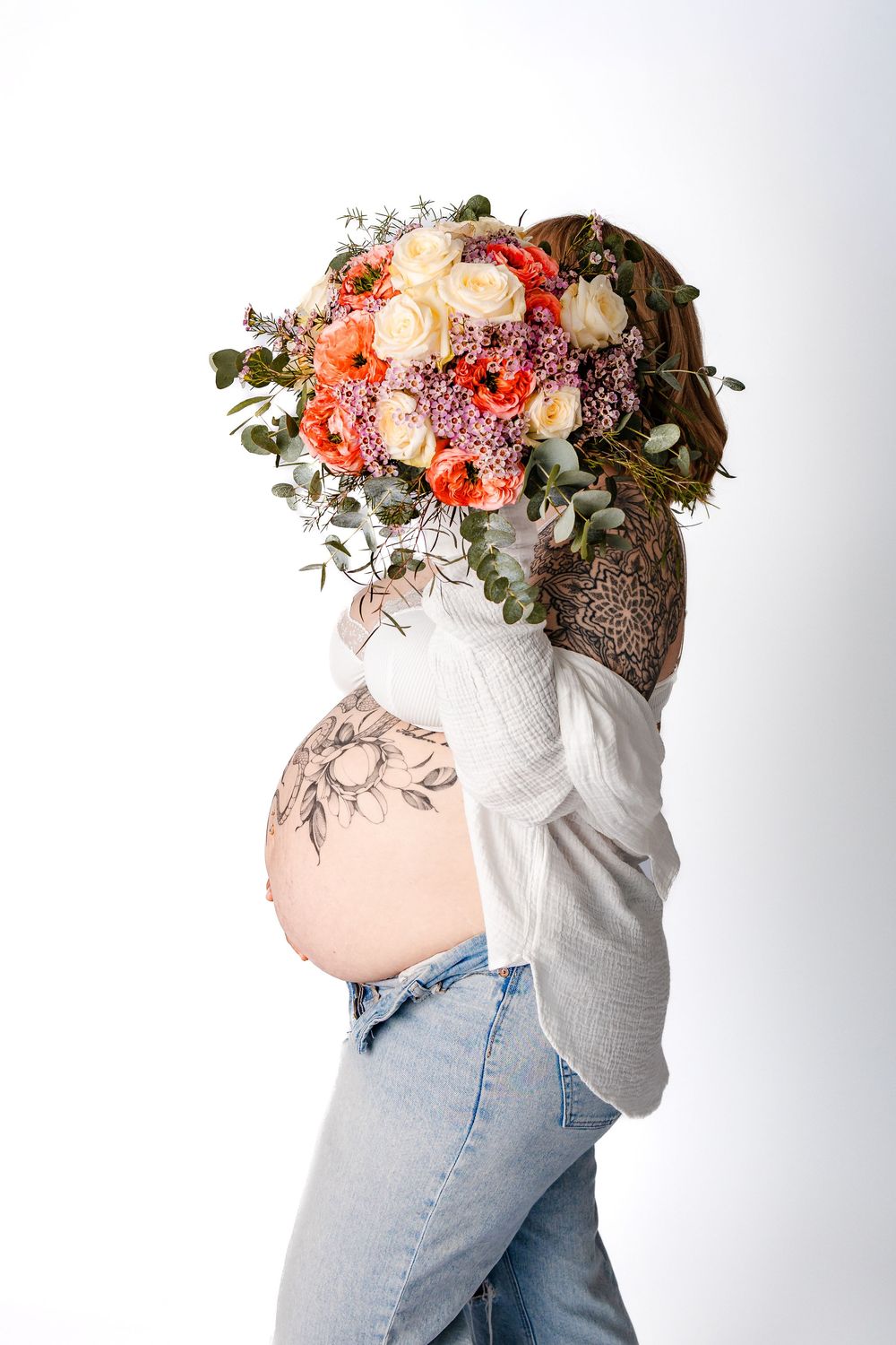 Babybauchshooting in jeans, einer bluse, einem Blumenstrauß und tattoos auf dem Bauch