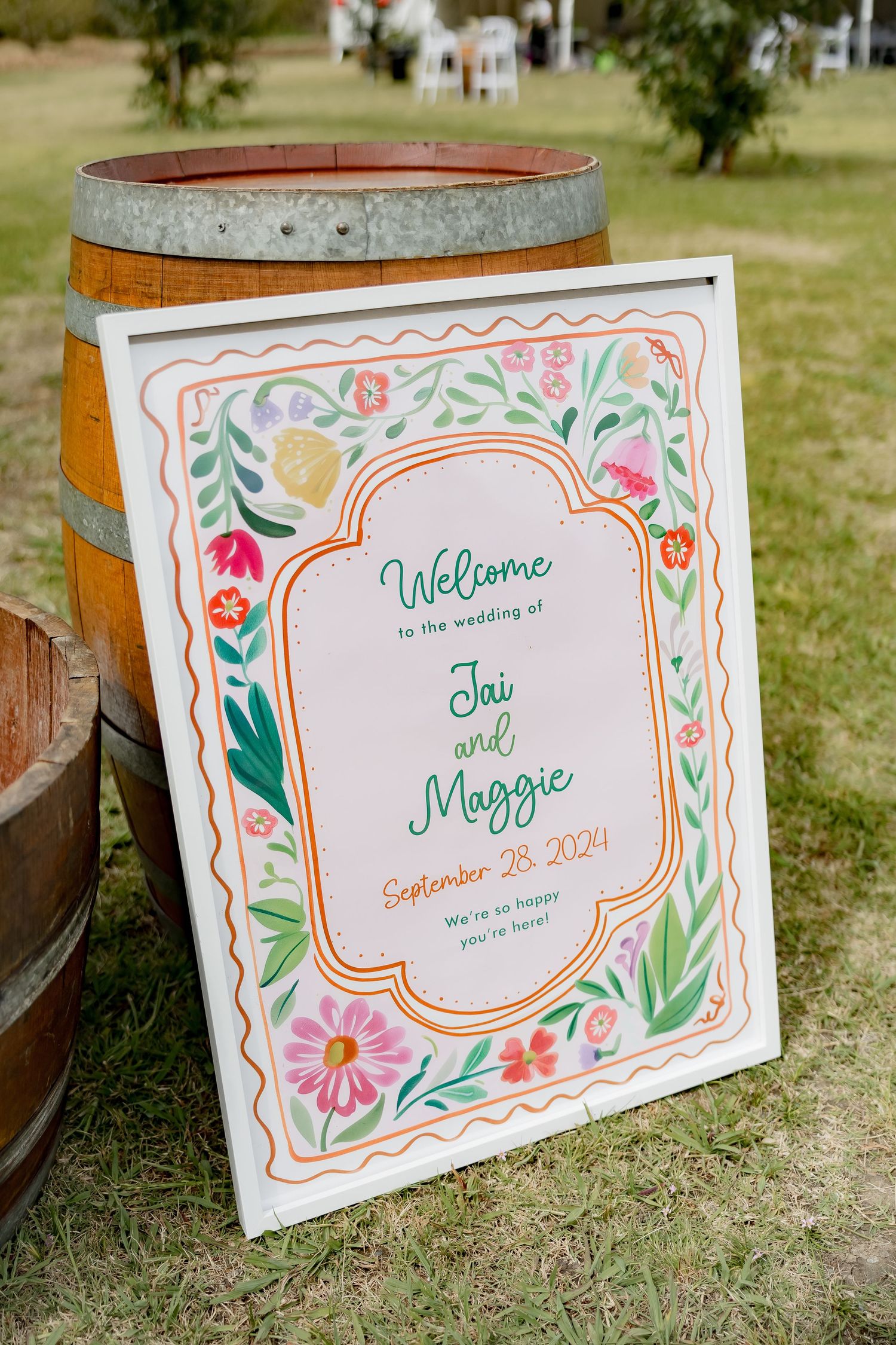 Colorful floral wedding welcome sign displayed on wooden barrel outdoors.