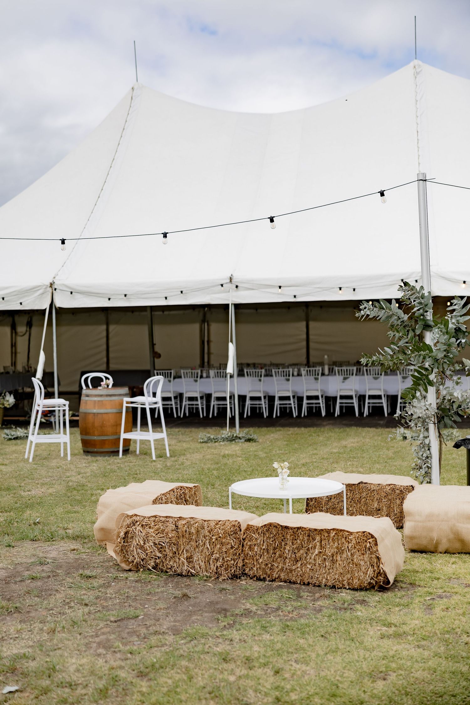 A white wedding tent set up outdoors with hay bale seating and string lights for a rustic reception setting.