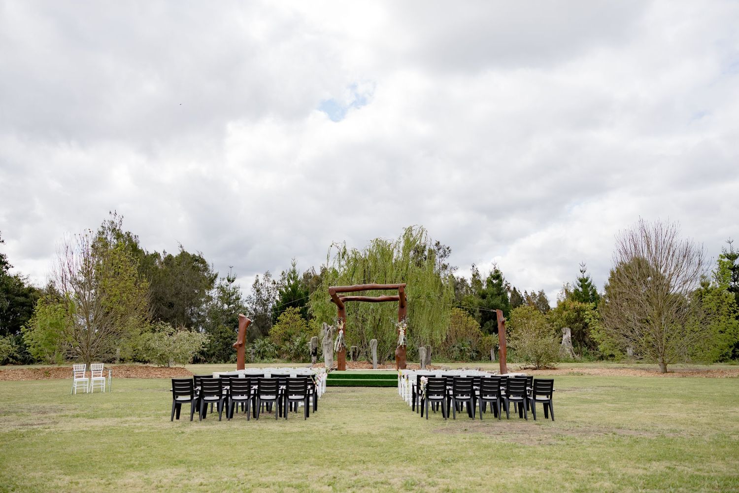 Outdoor wedding ceremony setup with black chairs and wooden arch in an open field.