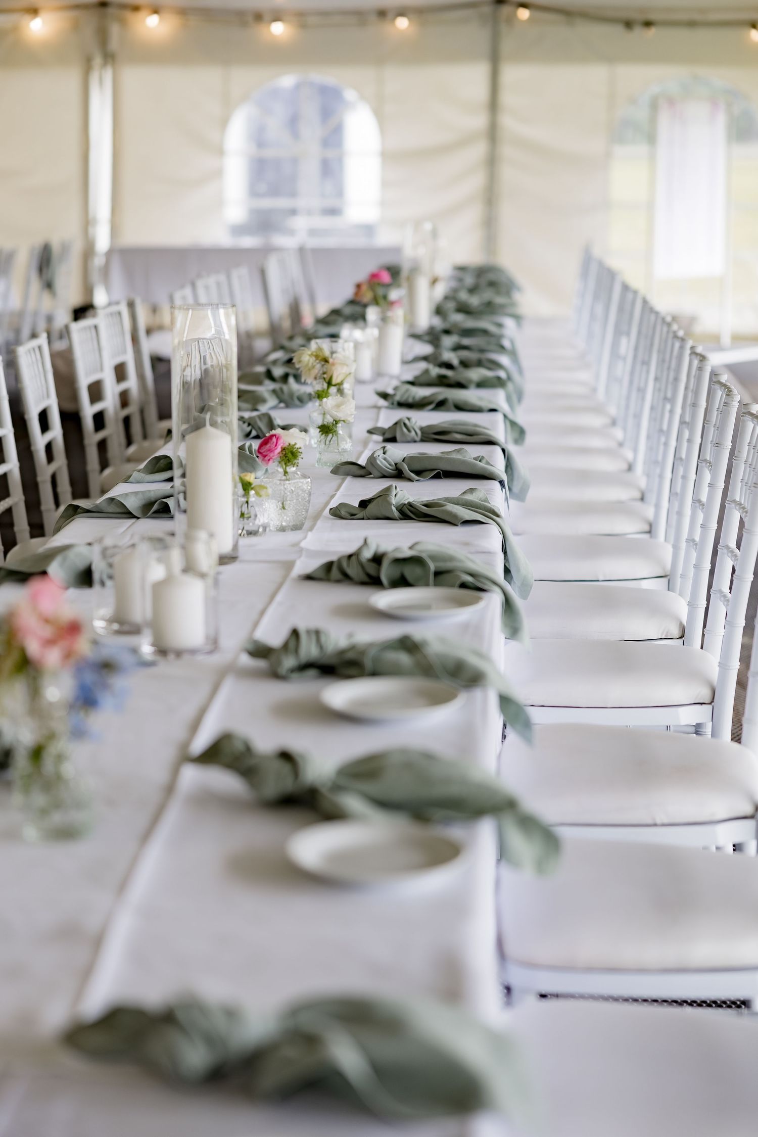 Elegant table setting with white chairs and sage green napkins in a wedding tent.