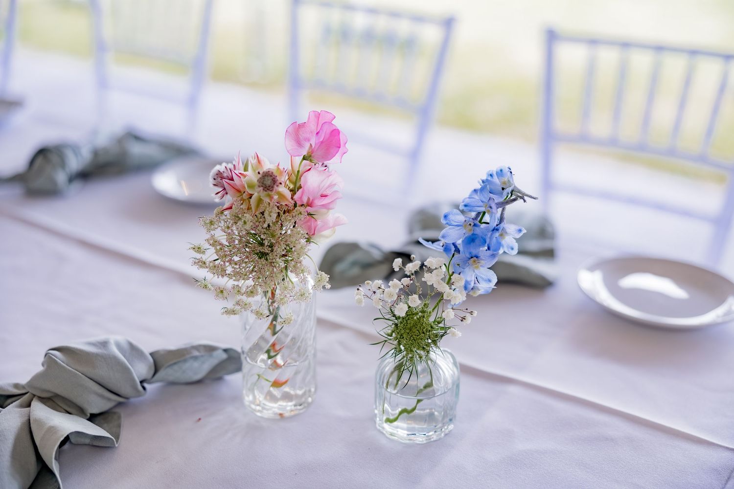 Delicate floral centerpieces with pink and blue flowers in small glass vases on wedding tables.