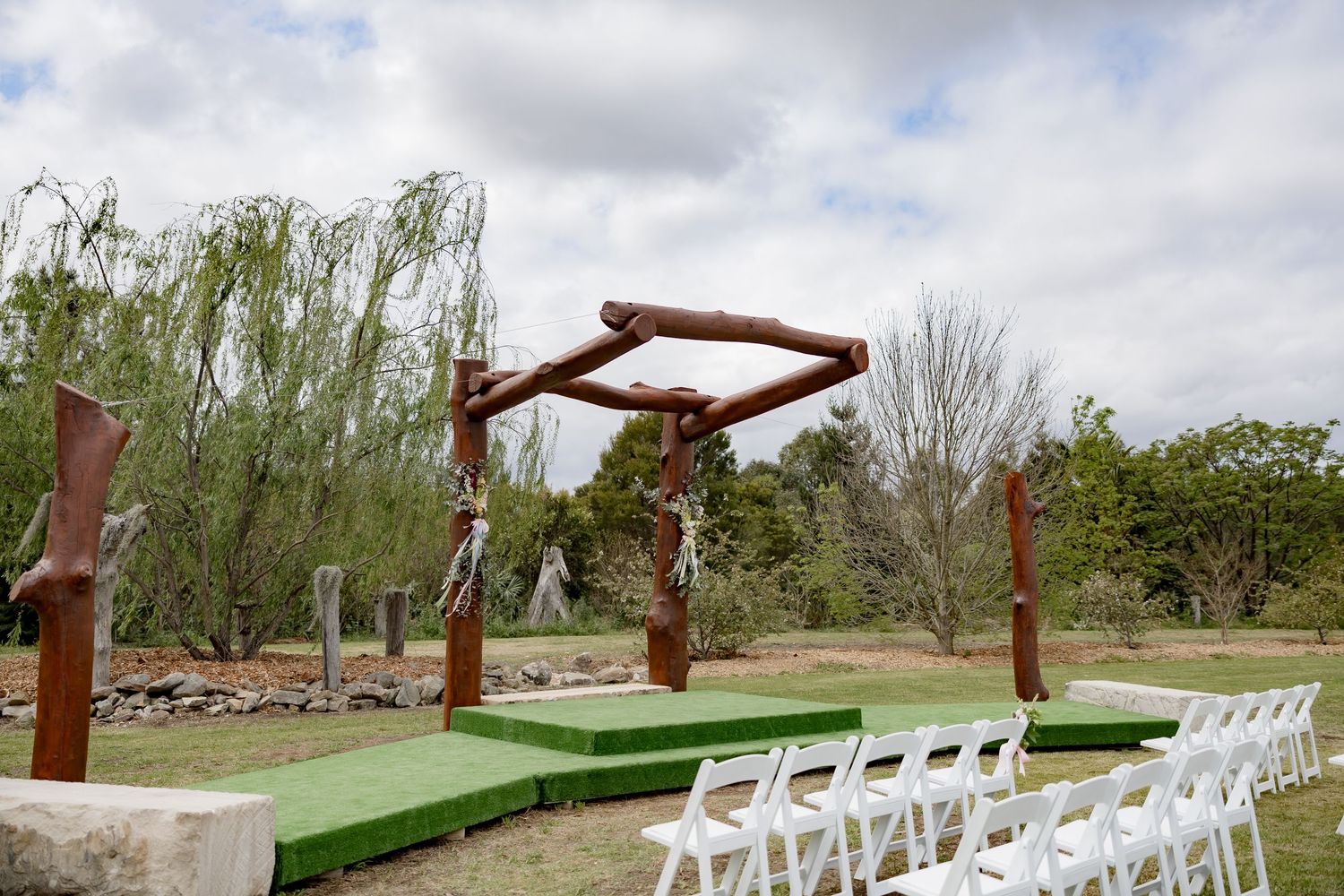Outdoor wedding ceremony setup with wooden arch and white chairs on grass.