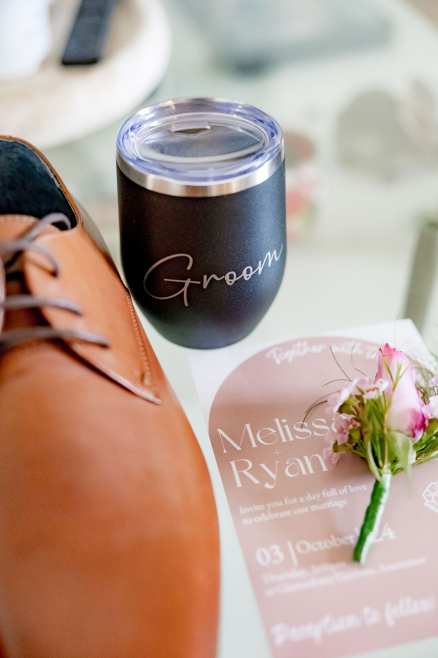 Wedding details including a black tumbler marked 'Groom', brown leather shoe, and pink floral boutonniere.