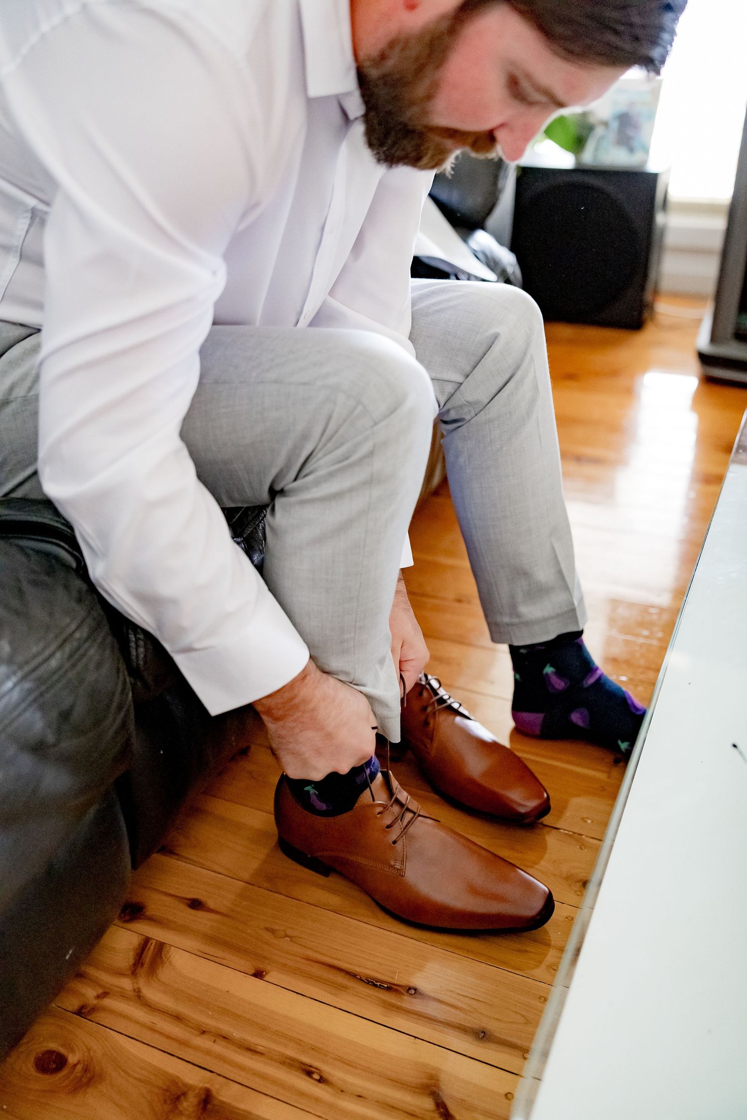 Person in white shirt and grey pants tying brown leather dress shoes on wooden floor.