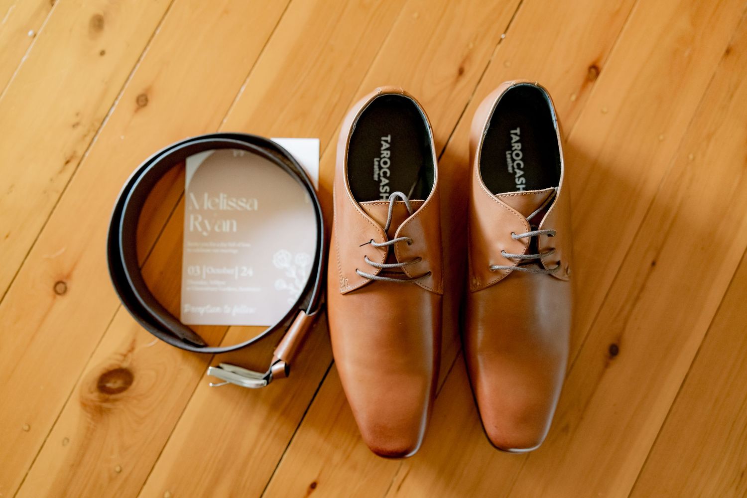 Brown leather dress shoes and belt arranged on wooden floor.