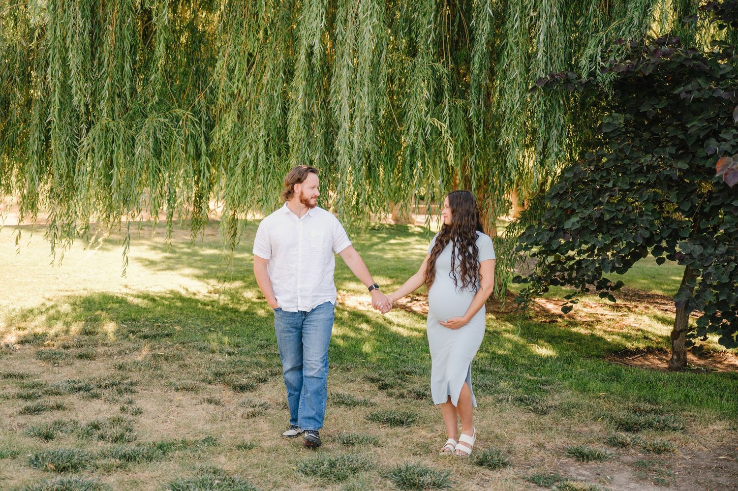 Couple walking hand in hand under weeping willow trees in a park setting.