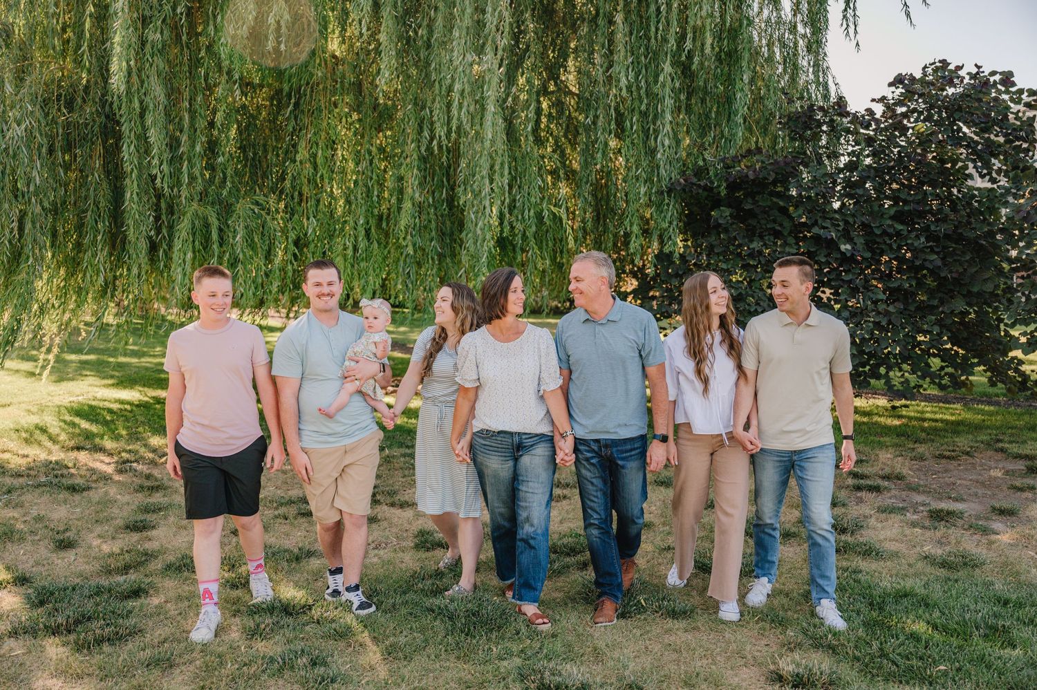 Group of people walking together under weeping willow trees in casual summer clothing.