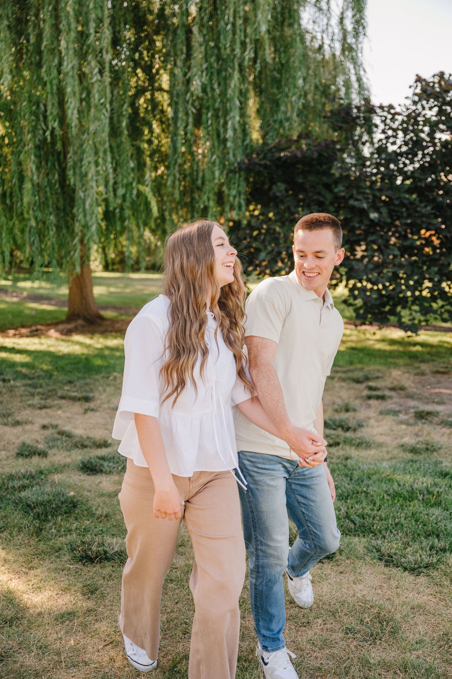 Couple sharing a joyful moment while walking under weeping willow trees in a park.