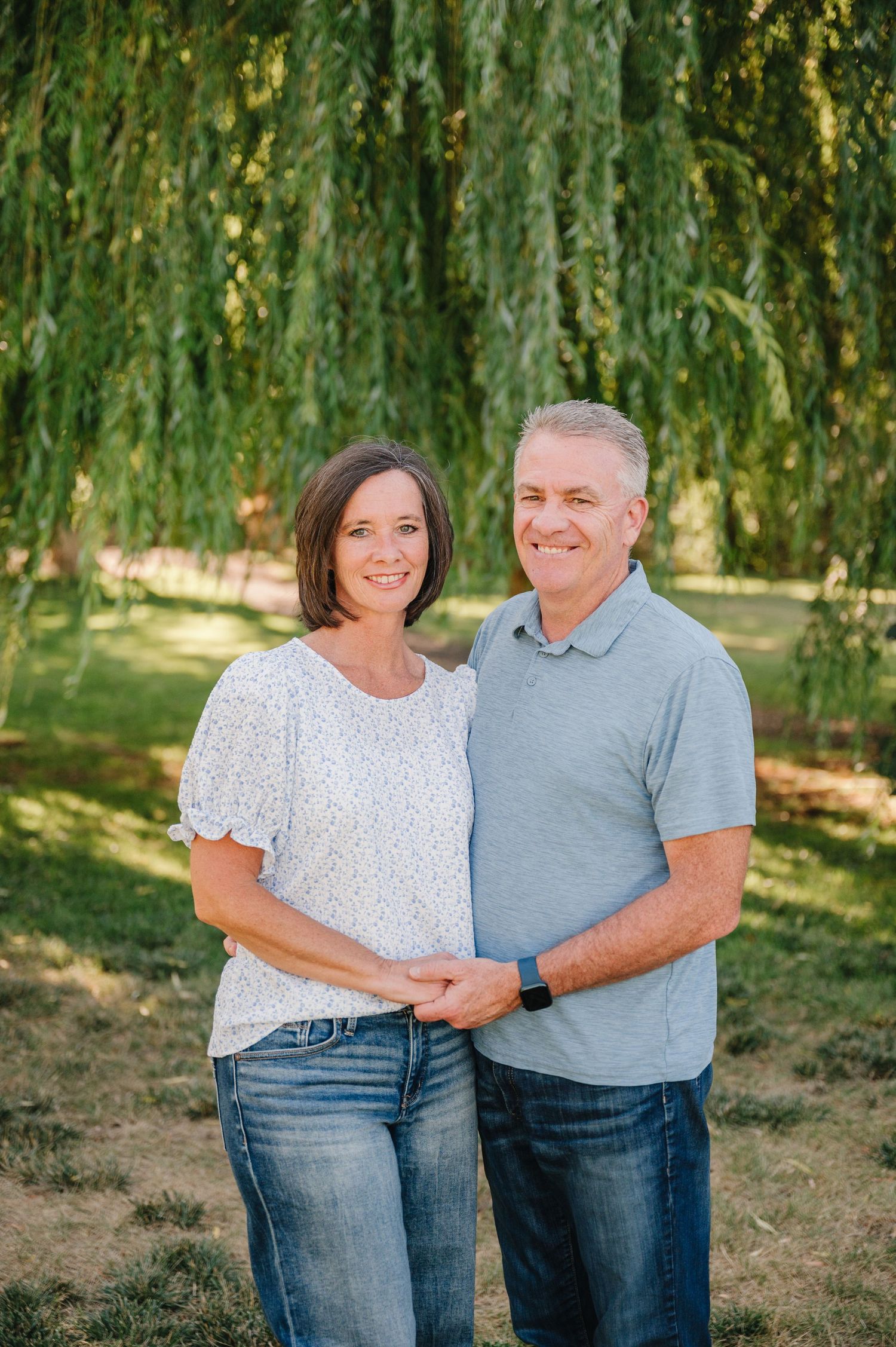 Couple in casual attire poses together under willow trees in summer.