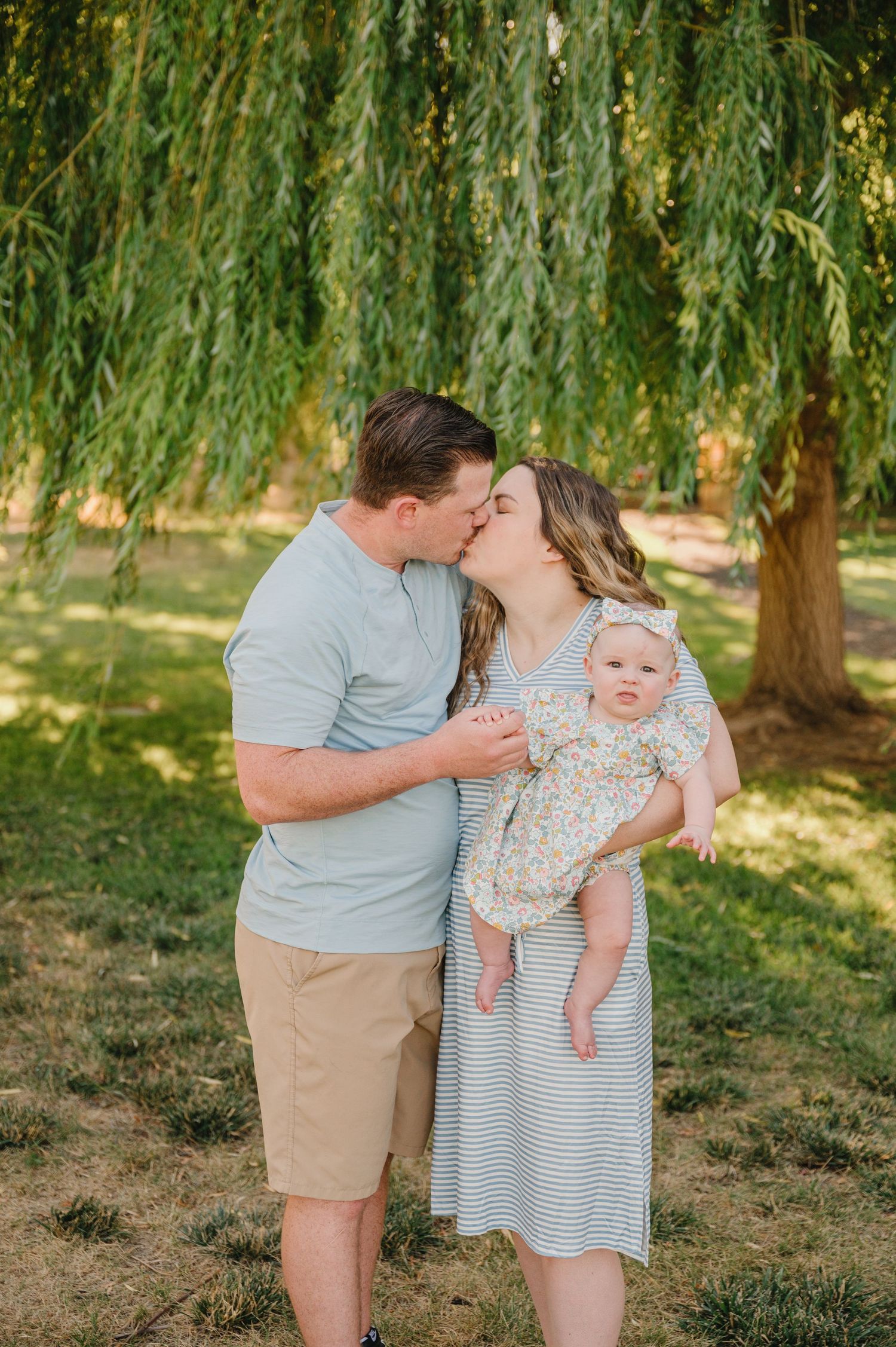 Family moment under willow trees with a baby in a floral outfit on a sunny day.