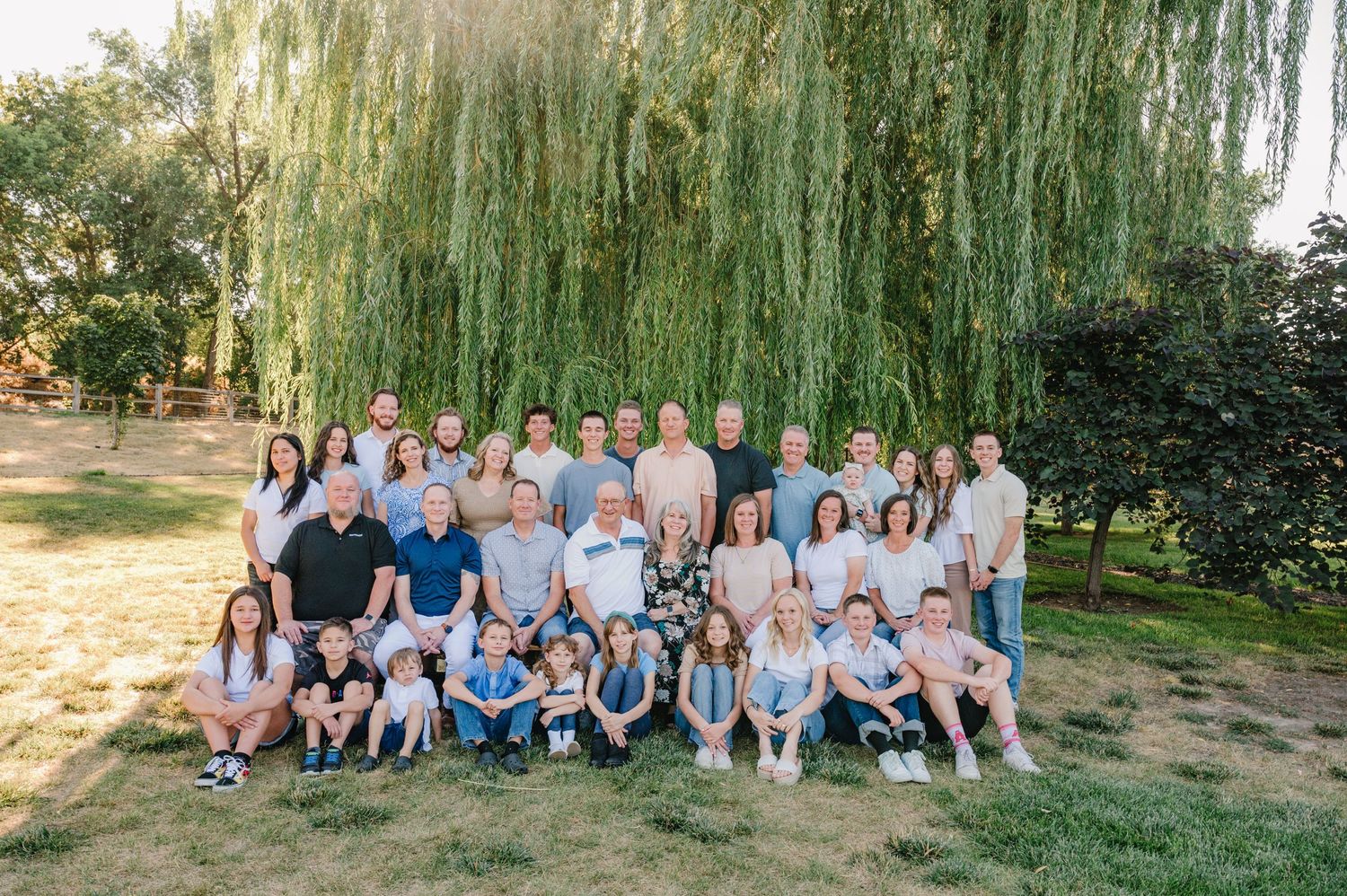 Extended family group photo with multiple generations seated on grass under willow trees.