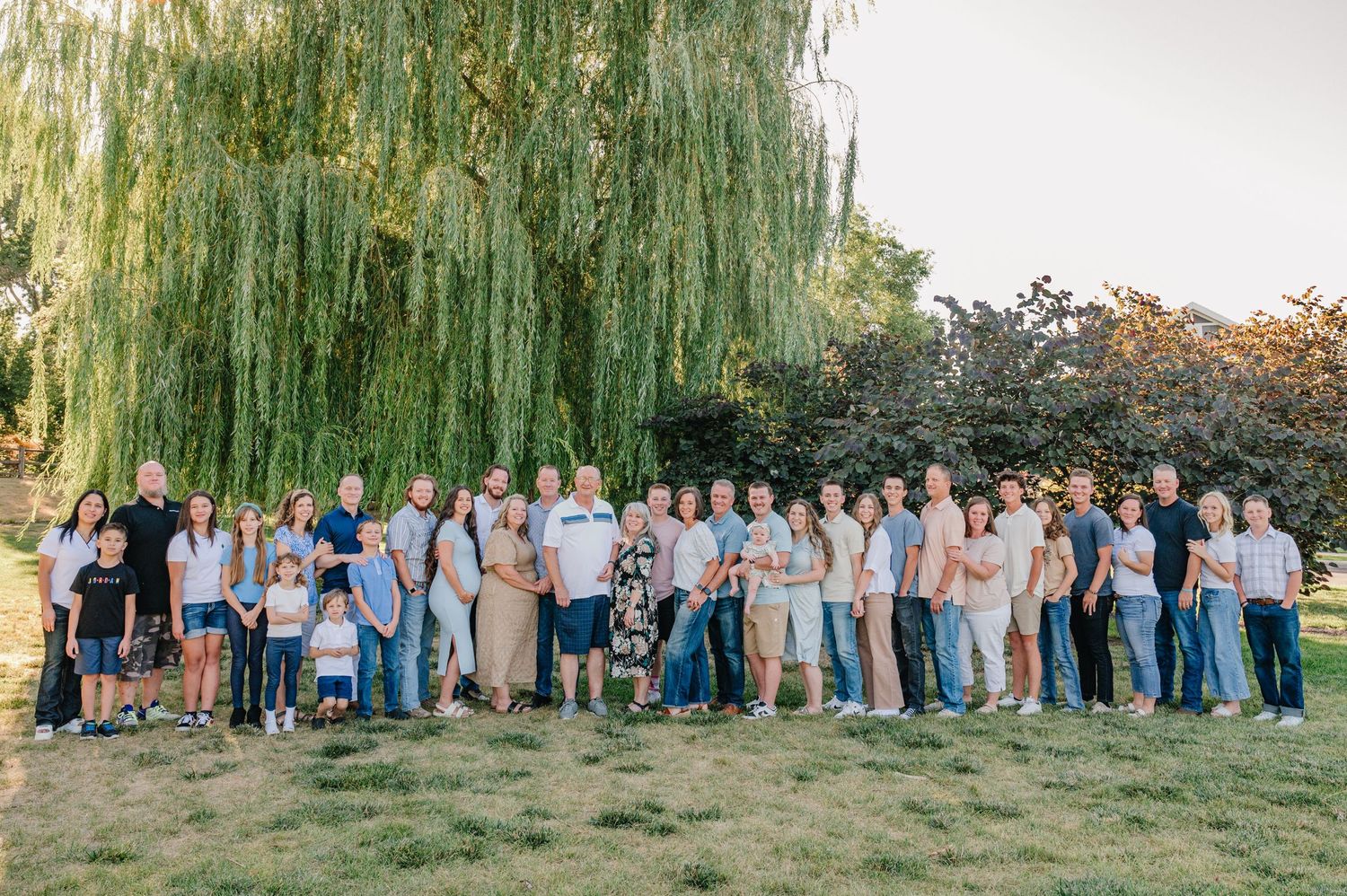 Large extended family gathering posed under weeping willow trees in coordinated blue and white outfits.