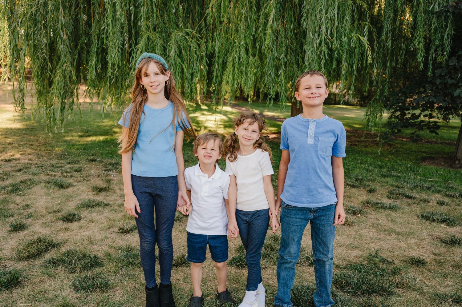 Four siblings pose together in casual clothing against a backdrop of drooping willow tree branches in a park setting.