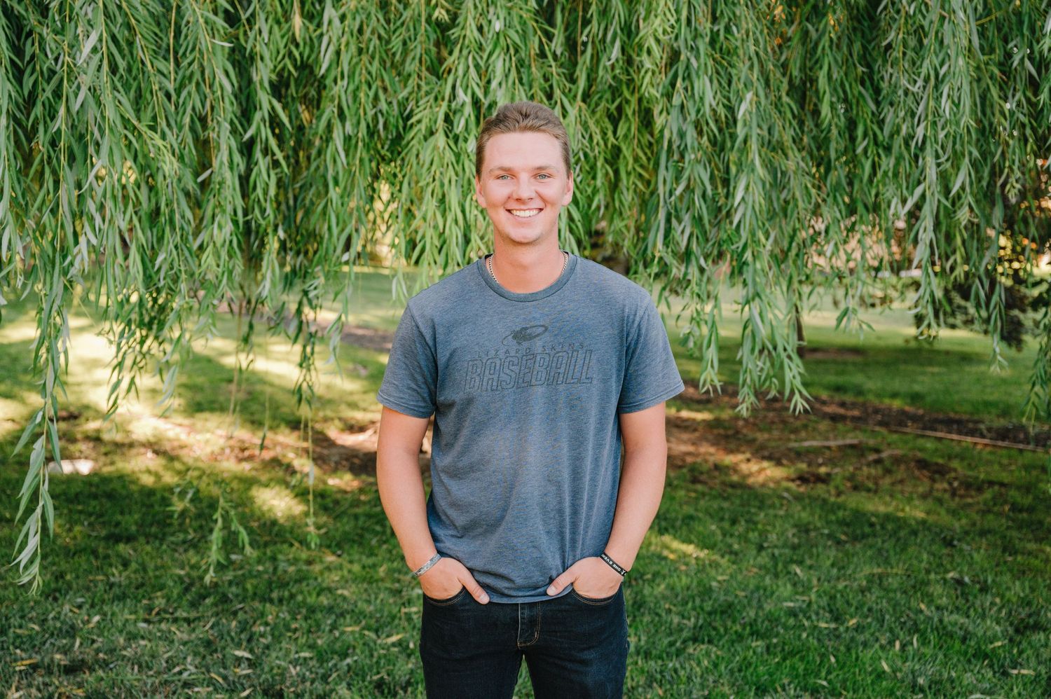 Person wearing grey t-shirt stands casually with hands in pockets under willow trees in summer.