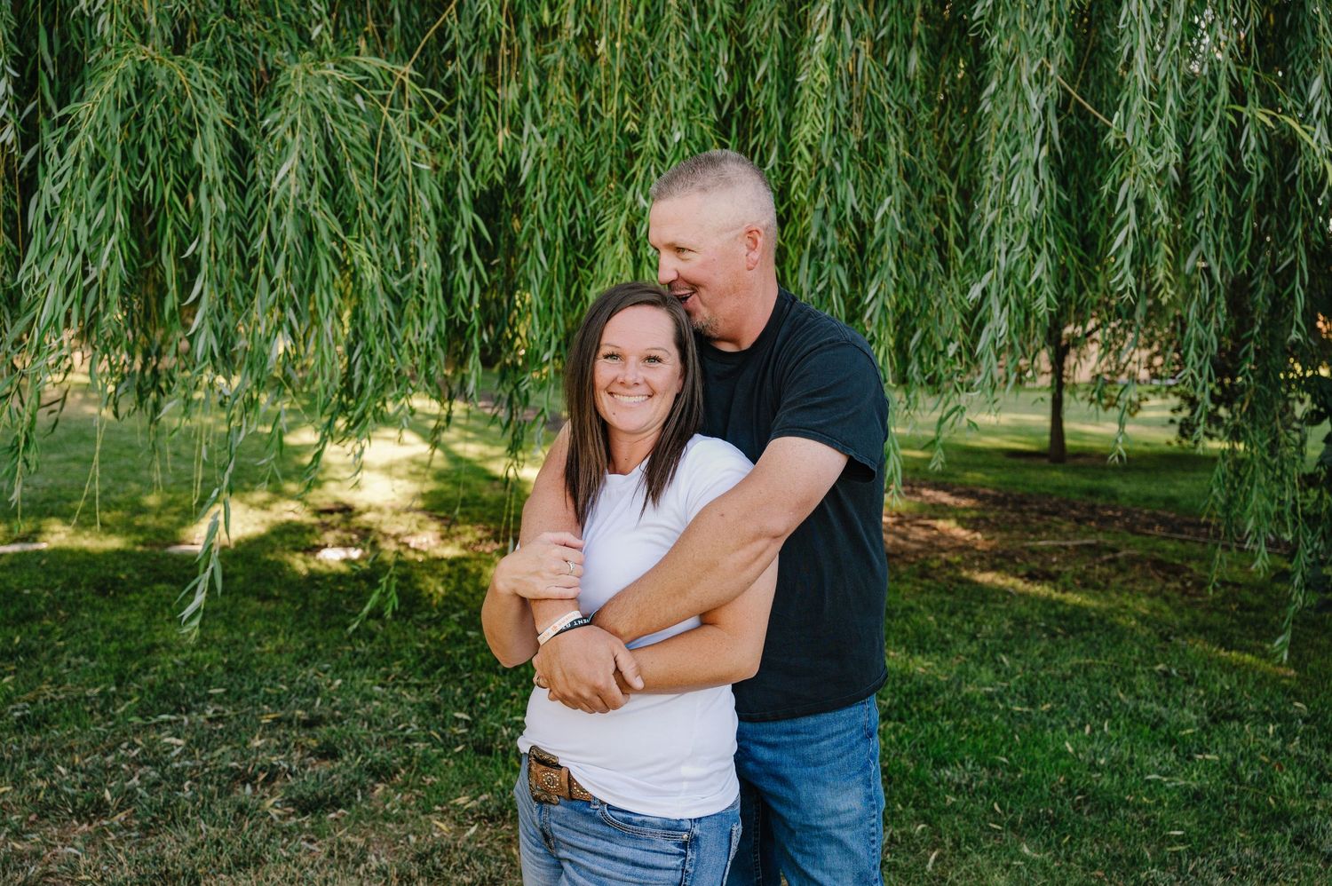 Two people embrace while standing in front of a beautiful weeping willow tree in a sunlit park setting.