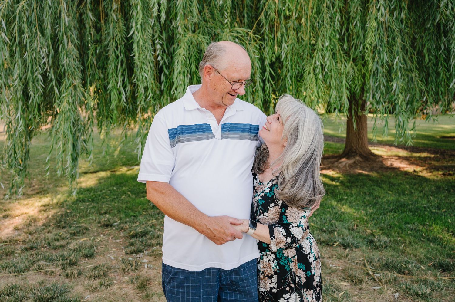 Couple shares a tender moment under weeping willow trees in a park.