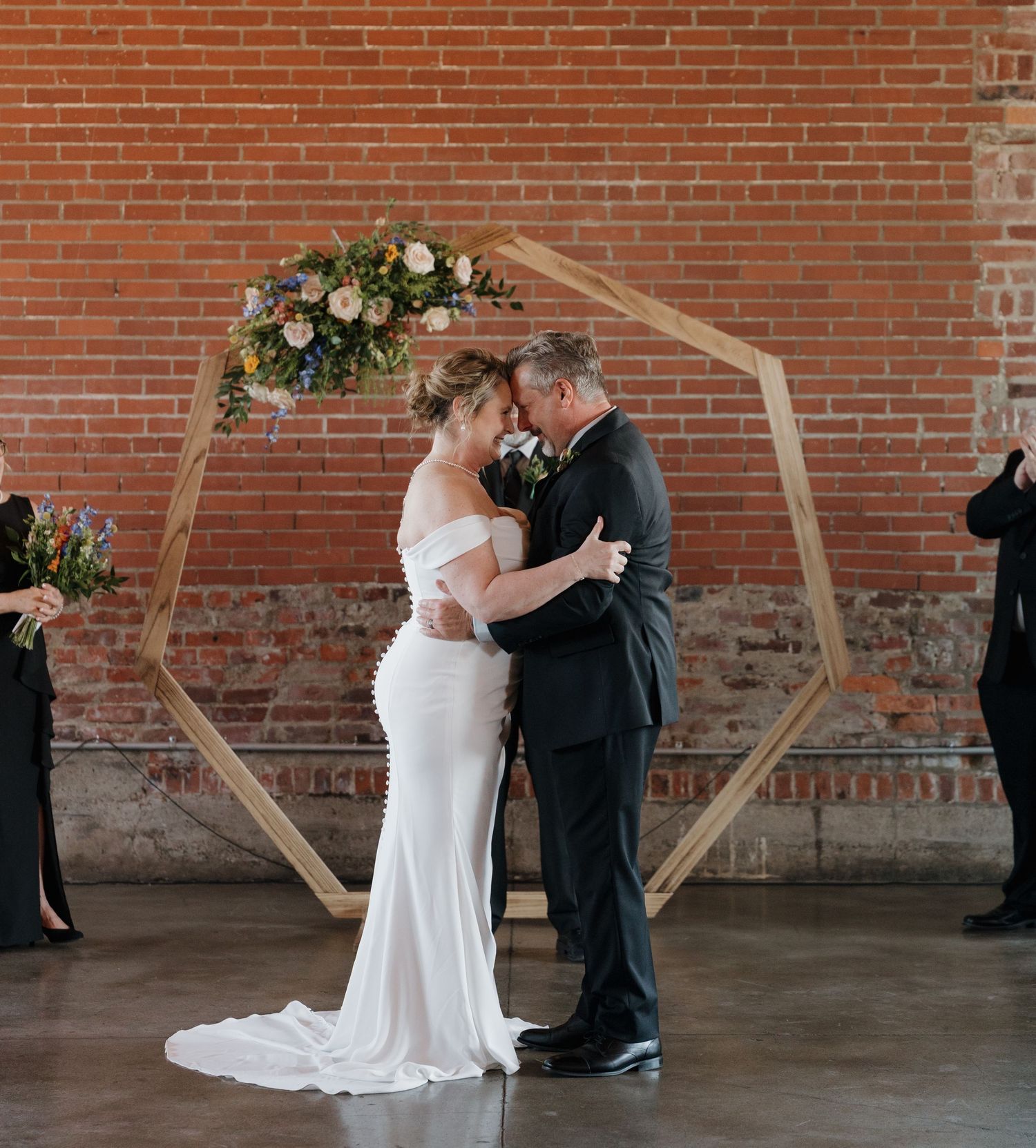 A couple shares their first kiss during an intimate wedding ceremony in front of a rustic wooden arch and brick wall.