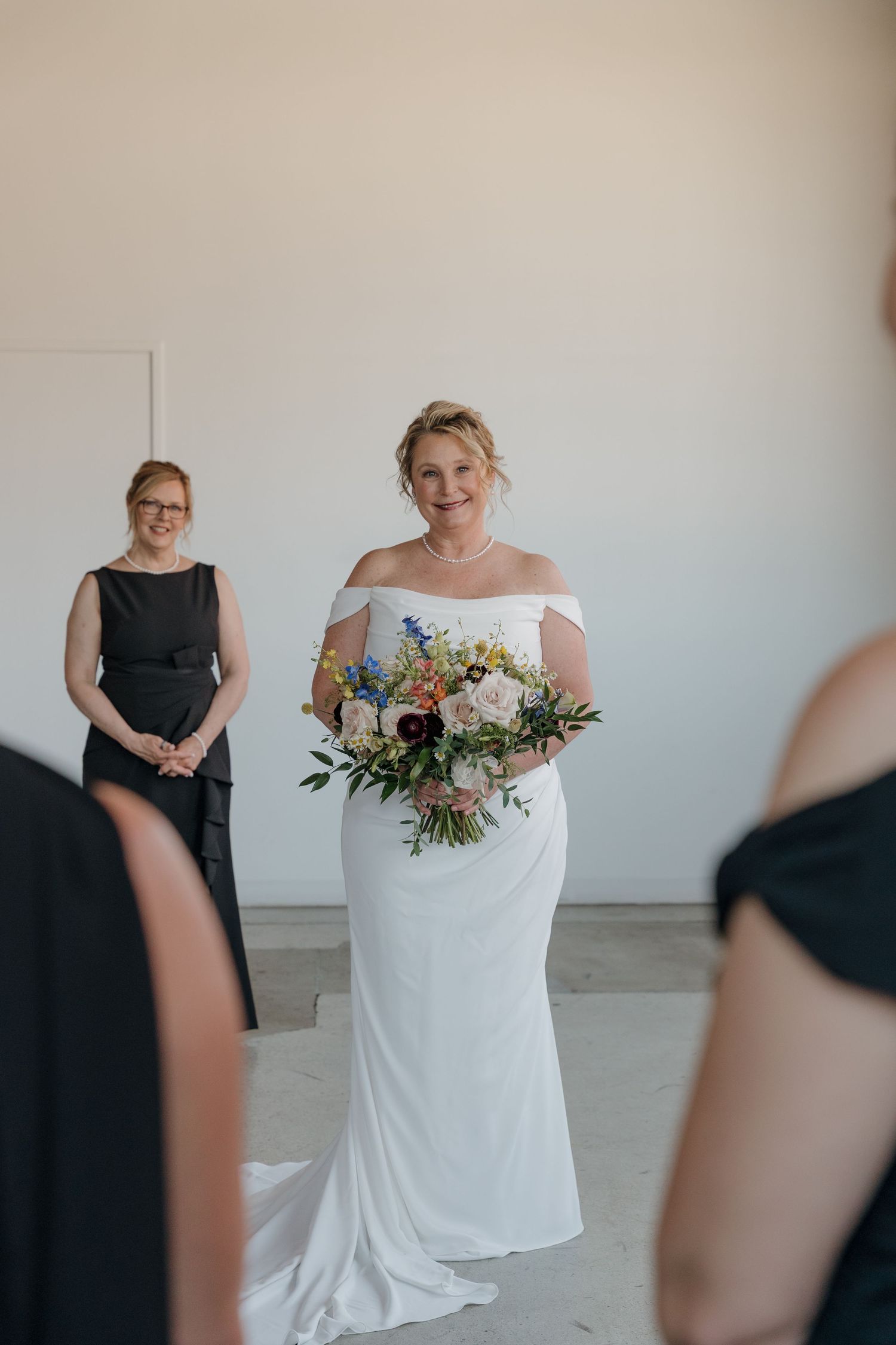 A bride in a white wedding dress holds a colorful floral bouquet while standing in a minimalist room with bridesmaids.