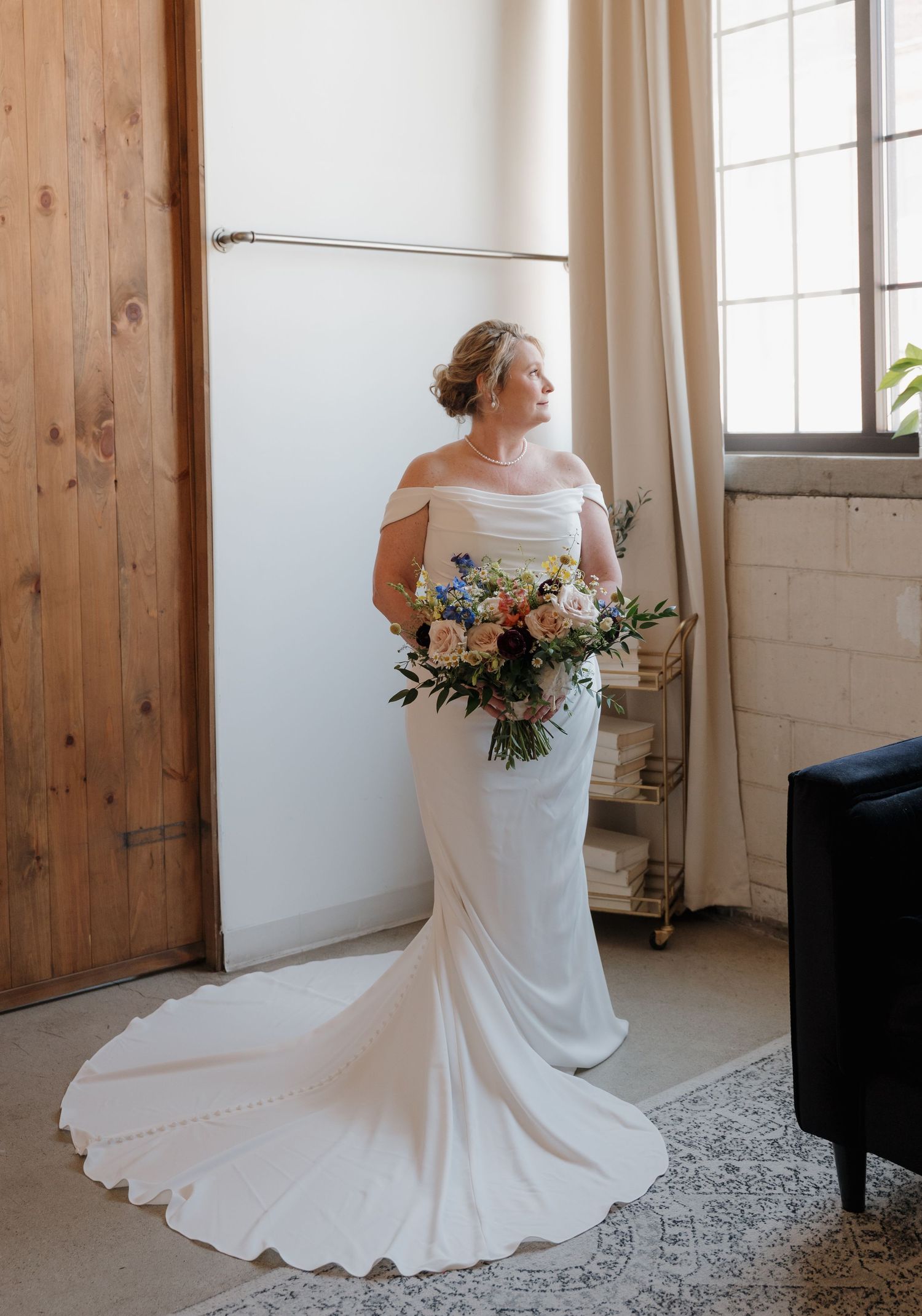 A bride in an elegant off-shoulder white wedding gown with a flowing train holds a romantic peach and white floral bouquet.