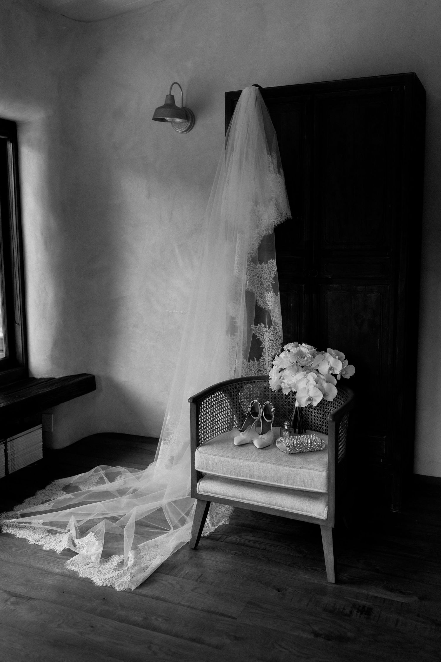 A vintage chair displays a flowing white wedding veil and bridal shoes next to fresh flowers in a moody black and white scene.