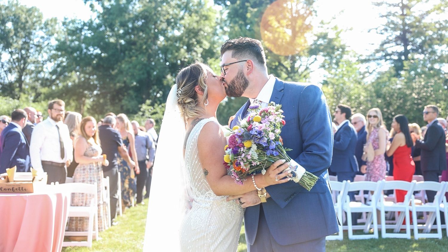A newlywed couple shares a kiss at an outdoor wedding ceremony with guests and white chairs in the background.