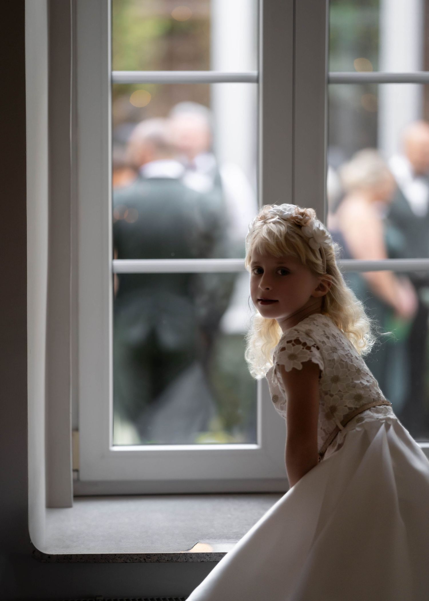 A young attendant in a white dress gazes out a sunlit window during a wedding celebration.