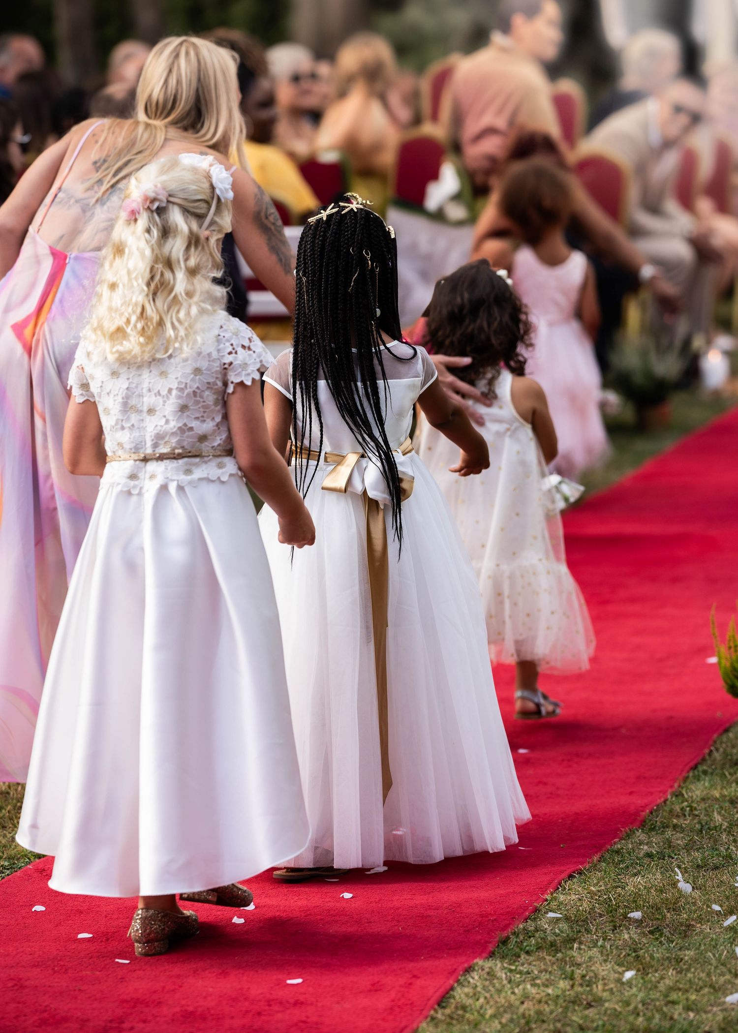 Young attendants in white dresses walking down a red carpet at an outdoor wedding.