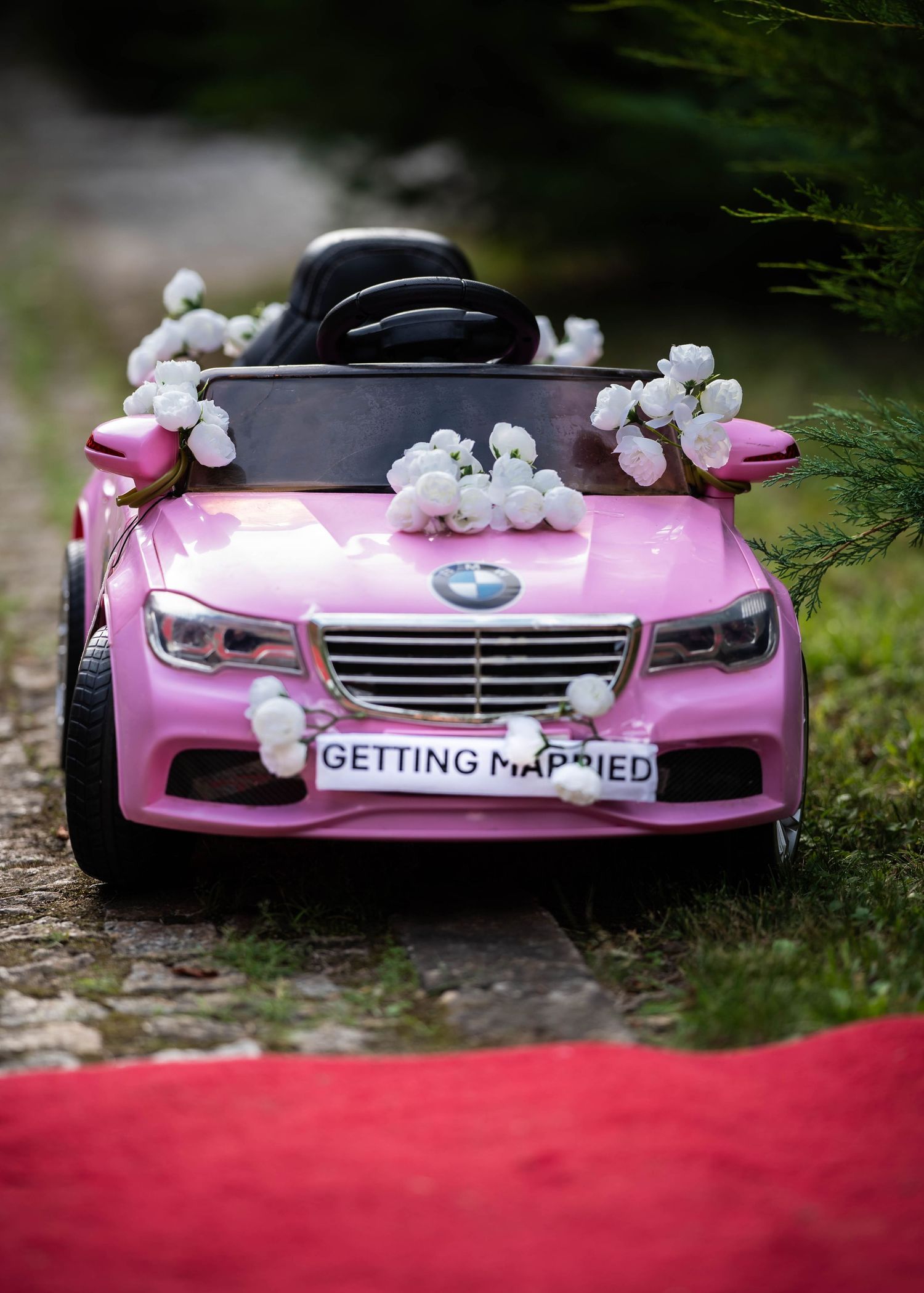 A pink toy car decorated with white flowers and a 'Getting Married' license plate.