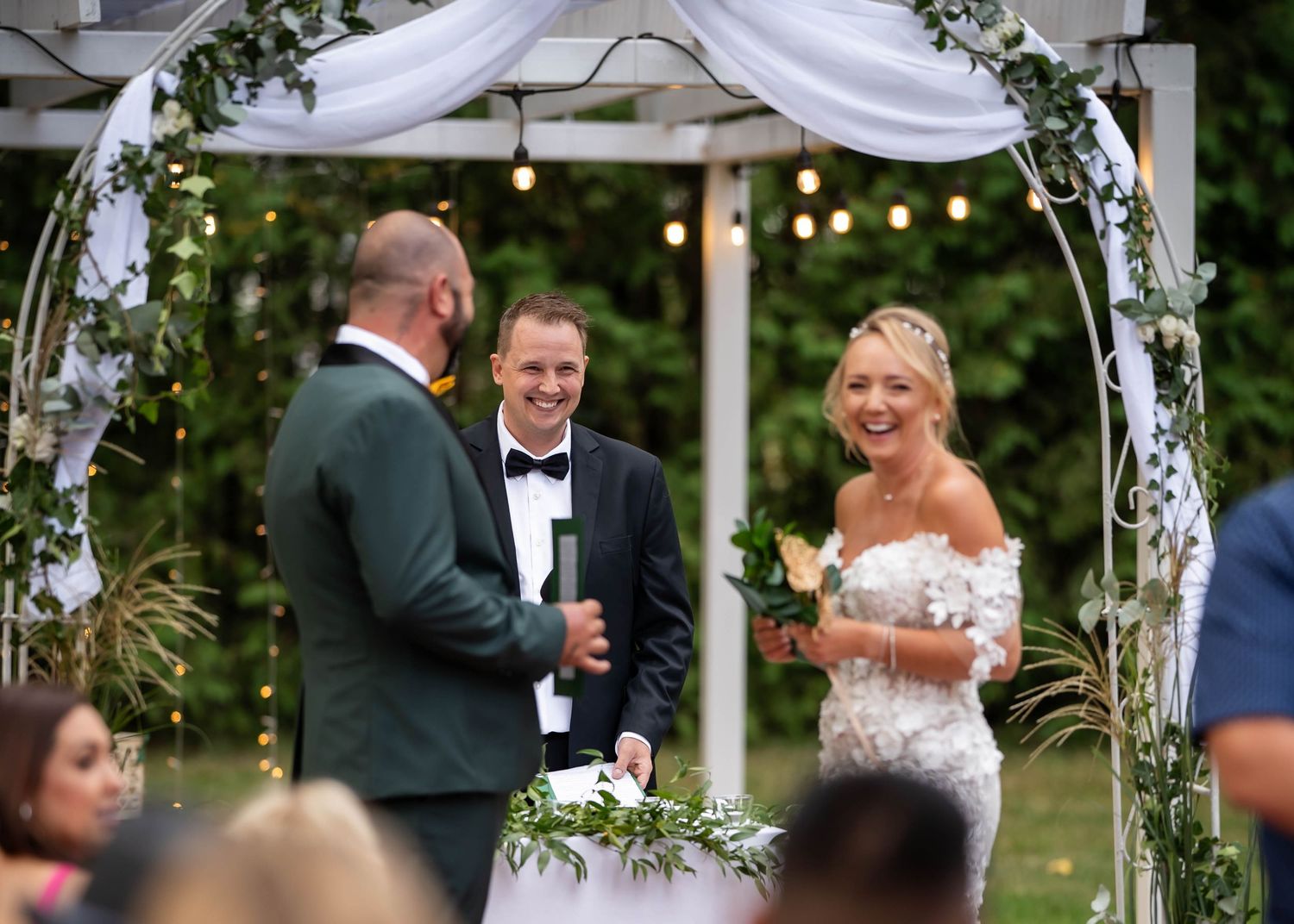 Outdoor wedding ceremony with couple exchanging vows under decorated arch with string lights.