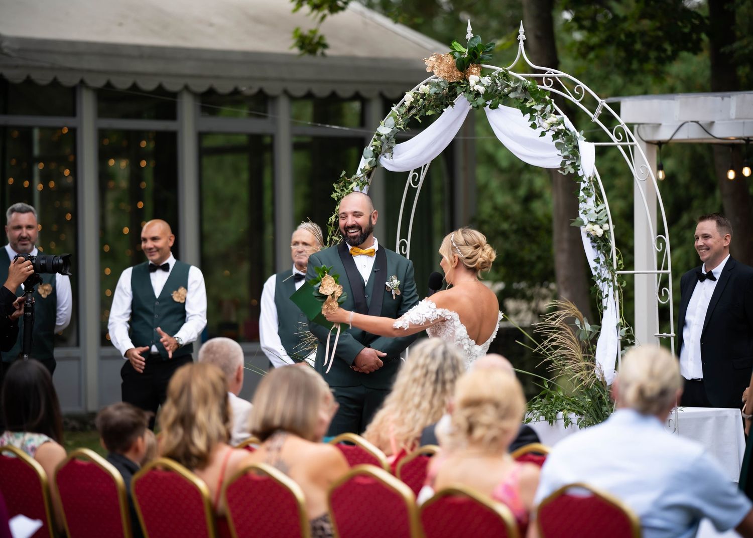 Wedding guests watch ceremony under decorated arch with white draping and greenery.