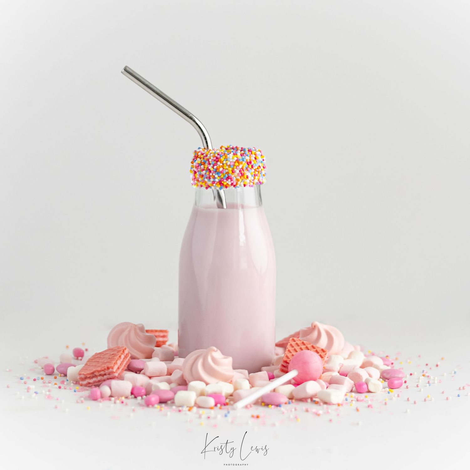 Pink milkshake in a small glass bottle with sprinkles on the rim of the bottle which is surrounded by sweets in shades of pink.