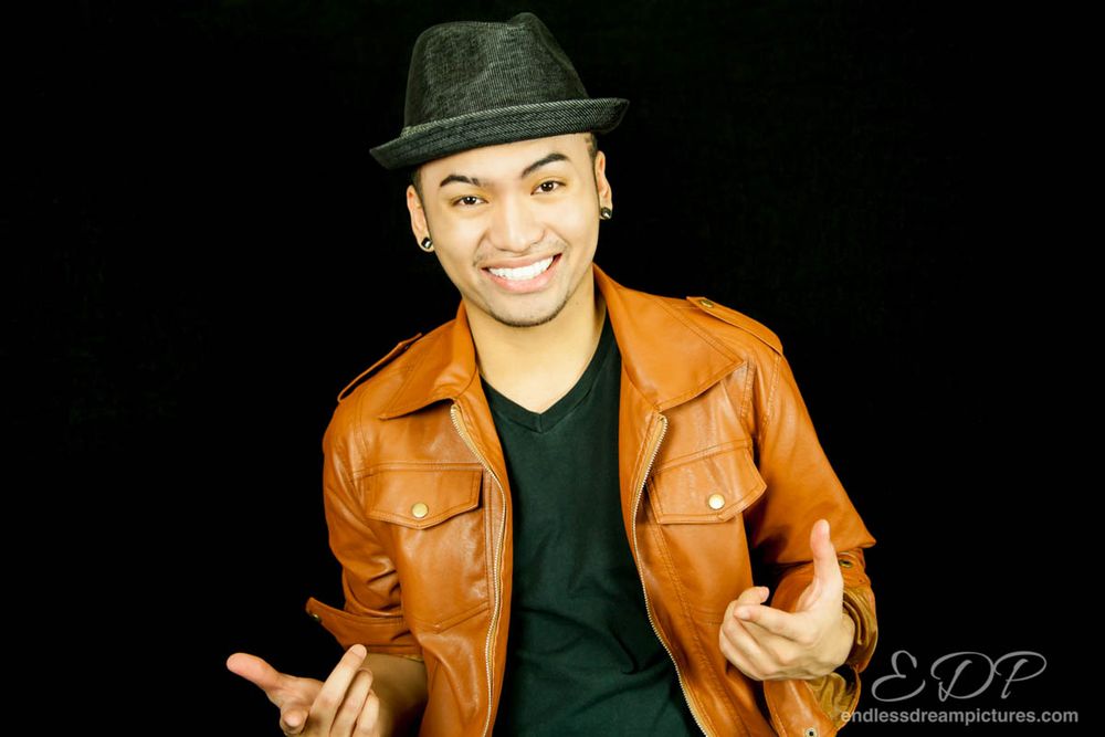 Stylish performer in orange leather jacket and black fedora gesturing against dark background.