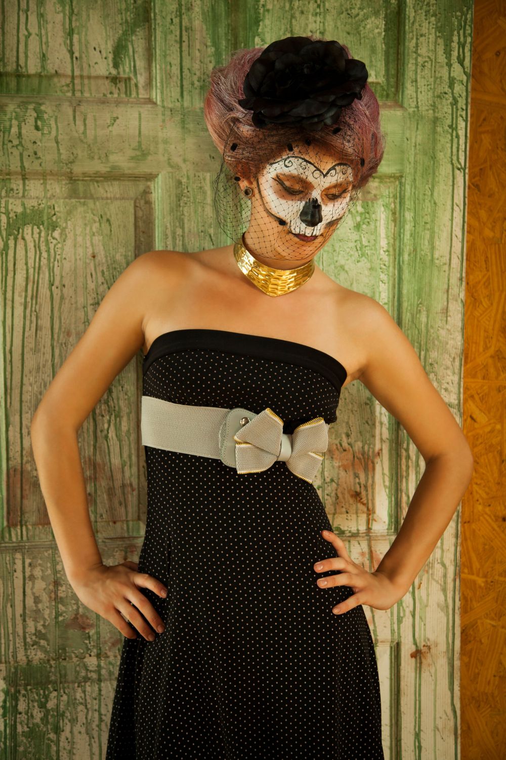Sugar skull makeup look with polka dot dress and statement bow belt against weathered green wall.