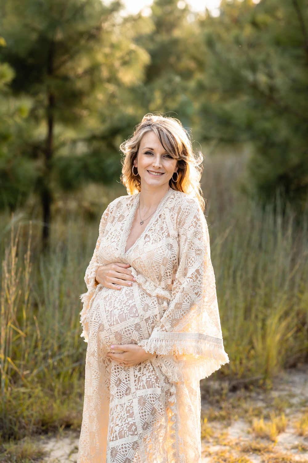 A sun-lit maternity portrait features a person in a flowing lace dress surrounded by golden light and tall grass.