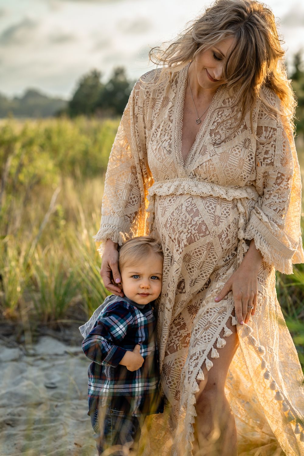 Intimate golden hour outdoor portrait showing maternal love with lace dress against natural field setting.