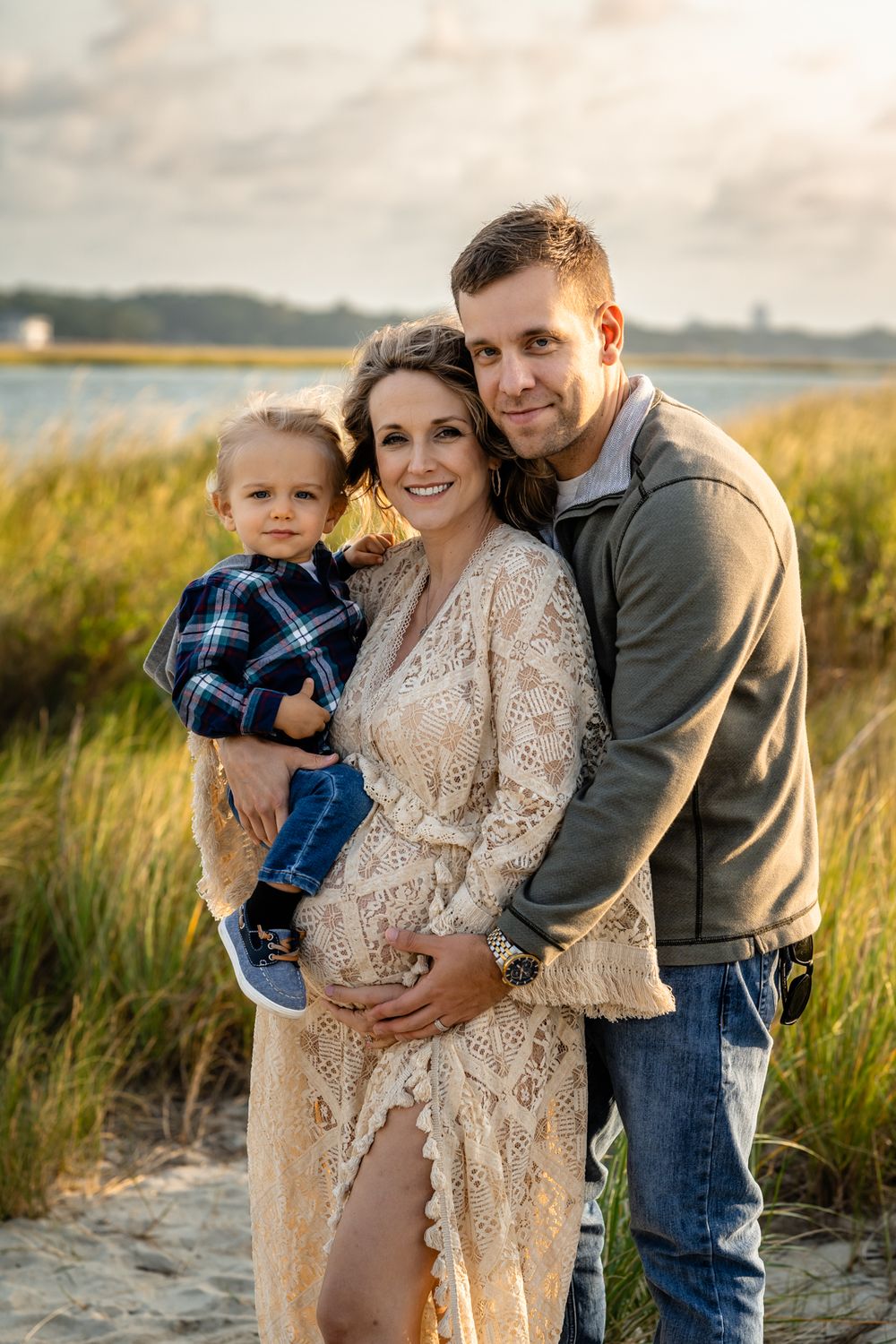 A heartwarming beach portrait of a growing family against golden light and coastal grass.
