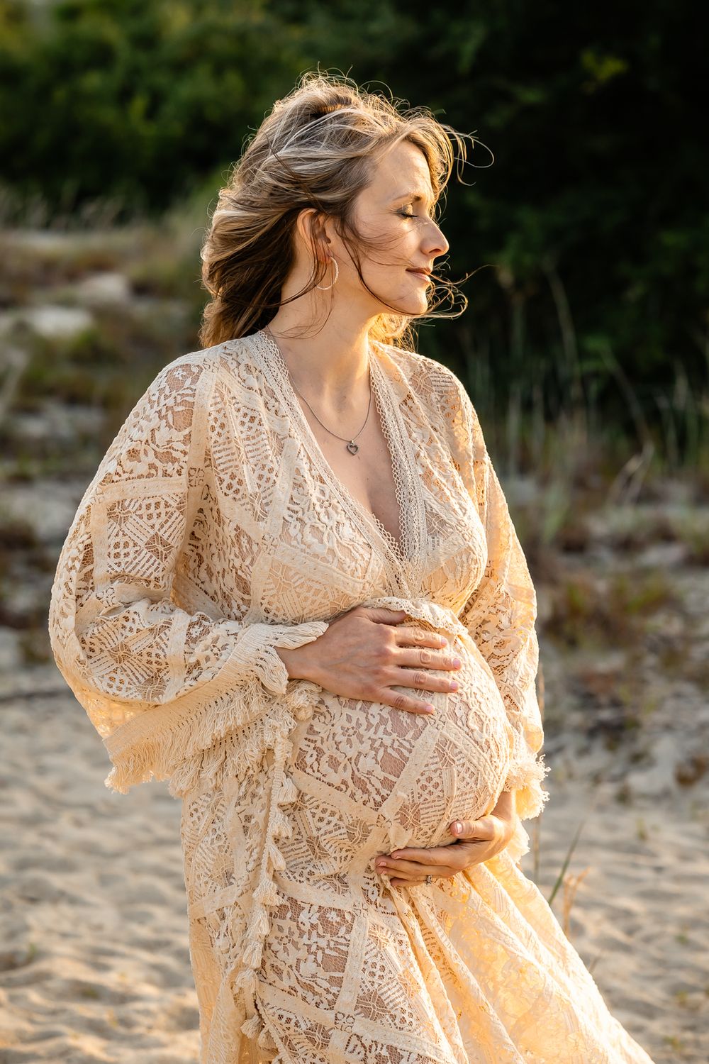 An expectant mother in a cream lace dress poses during golden hour on a beach.