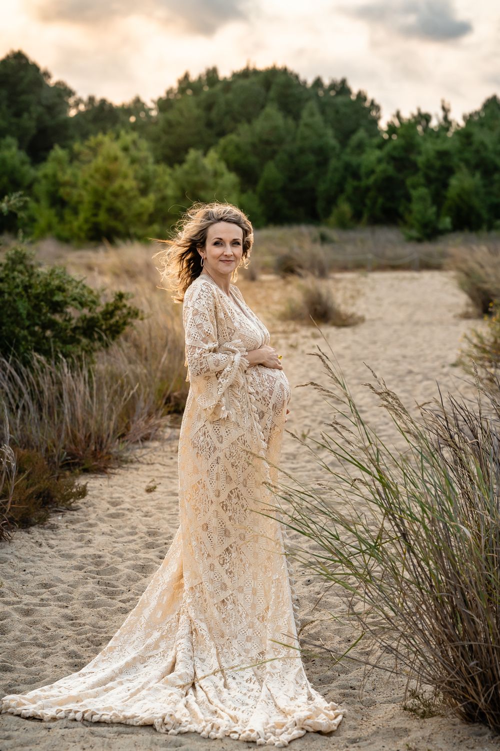 A dramatic full-length maternity portrait with flowing lace dress against coastal landscape.